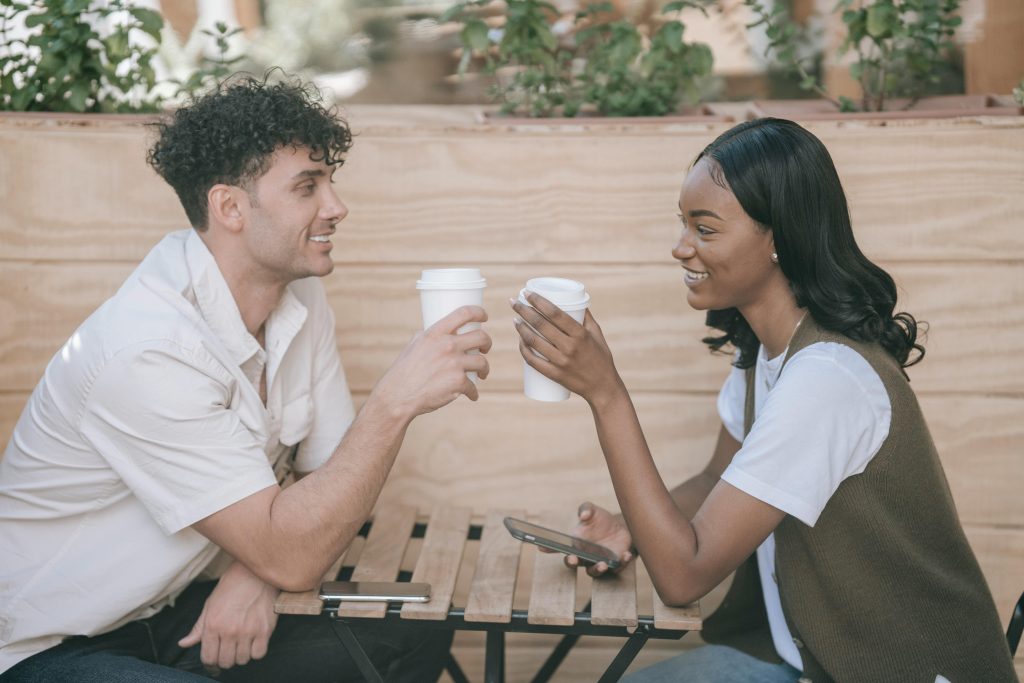 black woman and light-skinned man sitting and smiling at each other. Both people sitting across from each other holding white coffee cups.